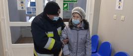 Zdjęcie przedstawia starszą kobietę ubraną w błękitną kurtkę oraz szary beret oraz mężczyznę w ubraniu strażackim. Mężczyzna trzyma kobietę pod pachę. Oboje znajdują się w przychodni lekarskiej.