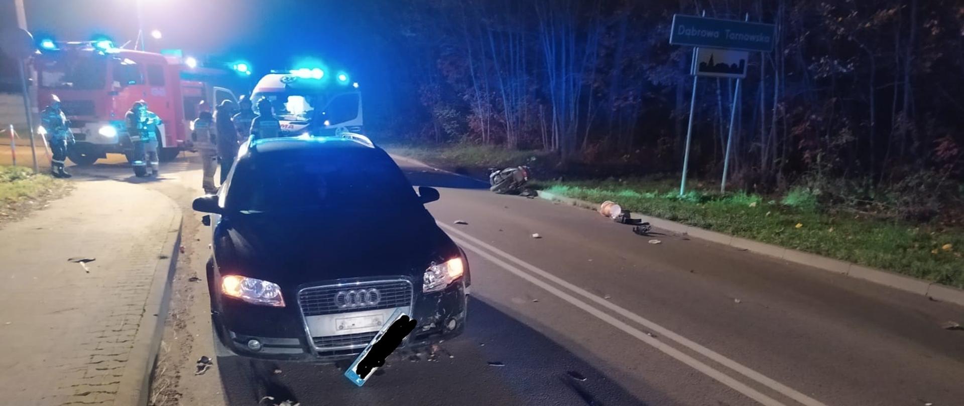 Na zdjęciu samochód biorący udział w zdarzeniu stoi na jezdni, obok leży motocykl, w oddali samochód straży pożarnej i zespół ratownictwa medycznego