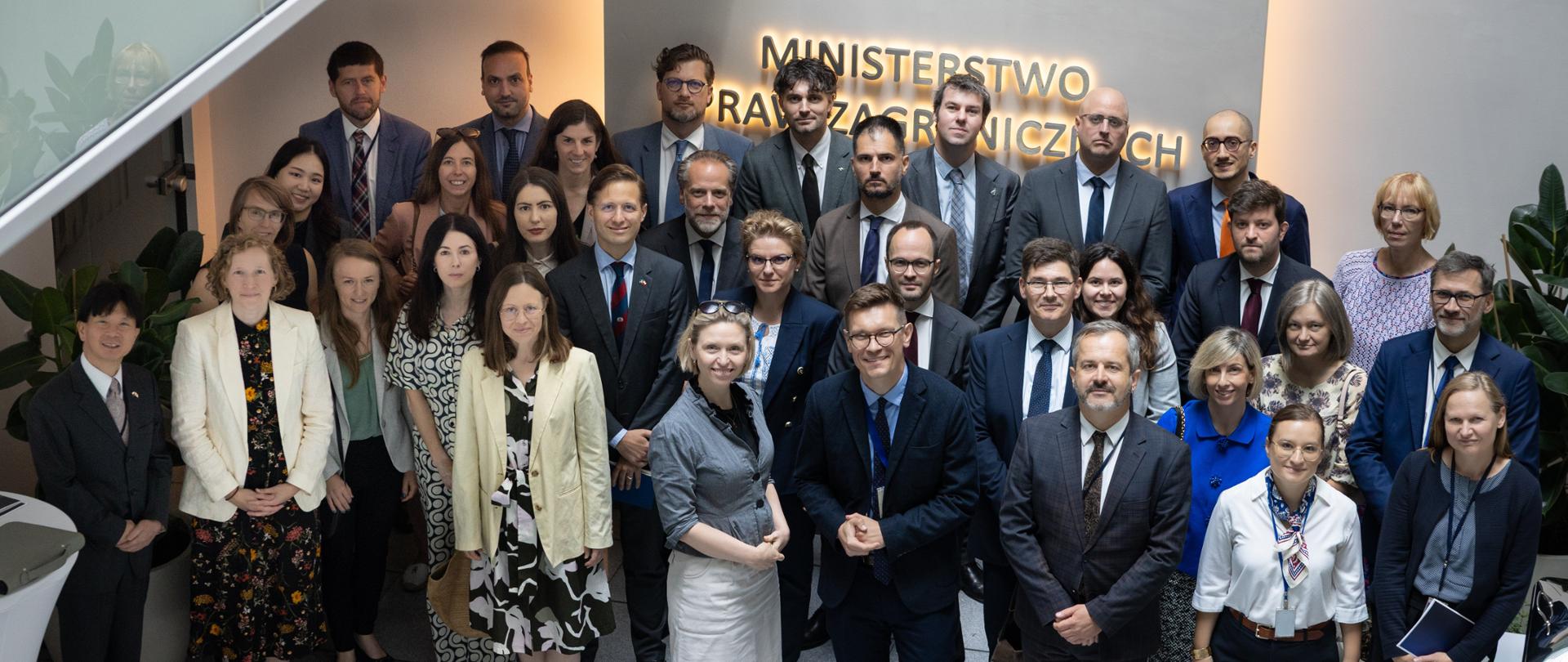 A group of a dozen or so people are standing in front of a sign reading ‘Ministry of Foreign Affairs’. They are looking towards the camera. 