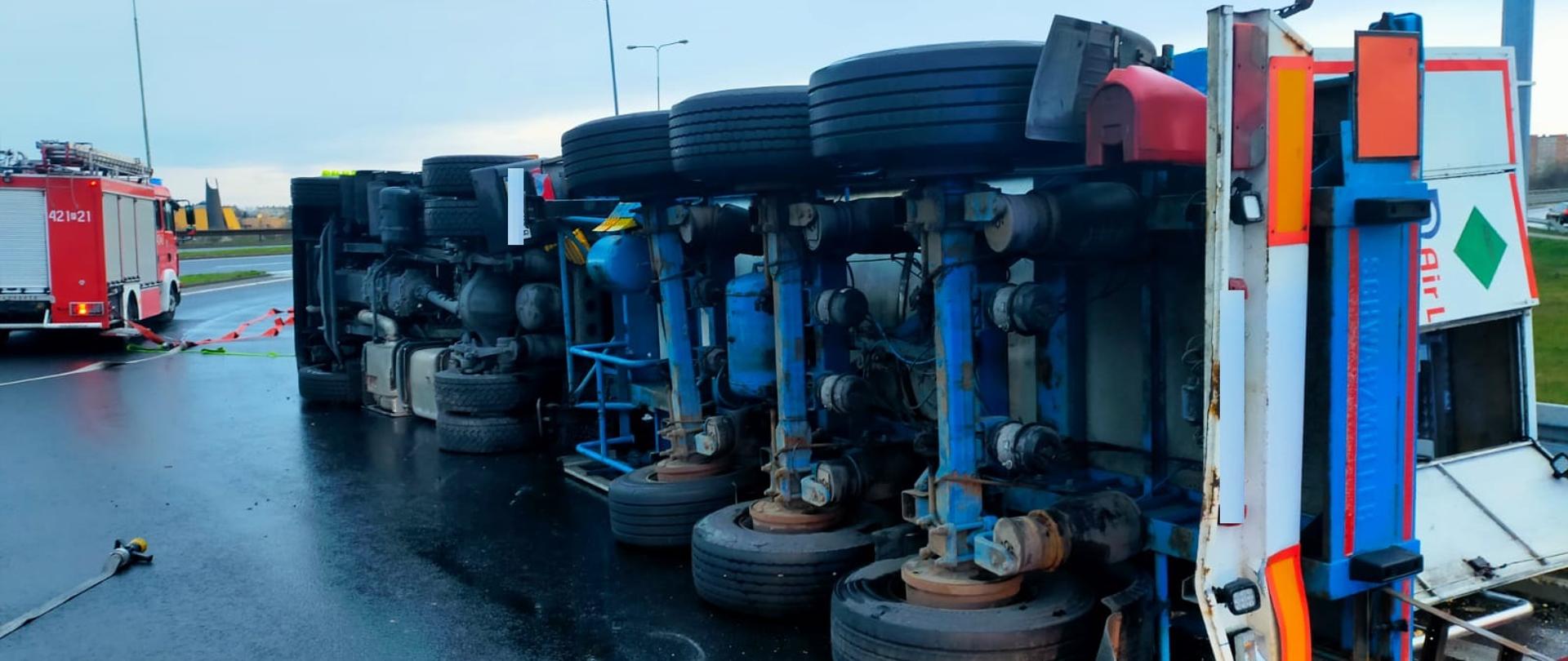 Mokra jezdnia. Leżąca na boku ciężarówka z czarnymi kołami. W tle czerwony wóz strażacki.