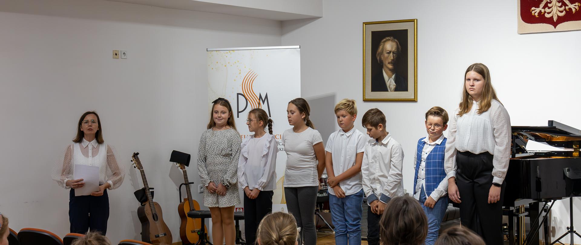 Międzynarodowy Dzień Muzyki 2023. Grupa uczniów, którzy przygotowali koncert oraz pani Agata Kopyto na scenie PSM w Głubczycach. W prawym górnym rogu widoczne godło Polski. W centrum zdjęcia za grupą uczniów widać baner reklamowy szkoły z okazji 50-lecia PSM. Na pierwszym planie u dołu zdjęcia widać publiczność.