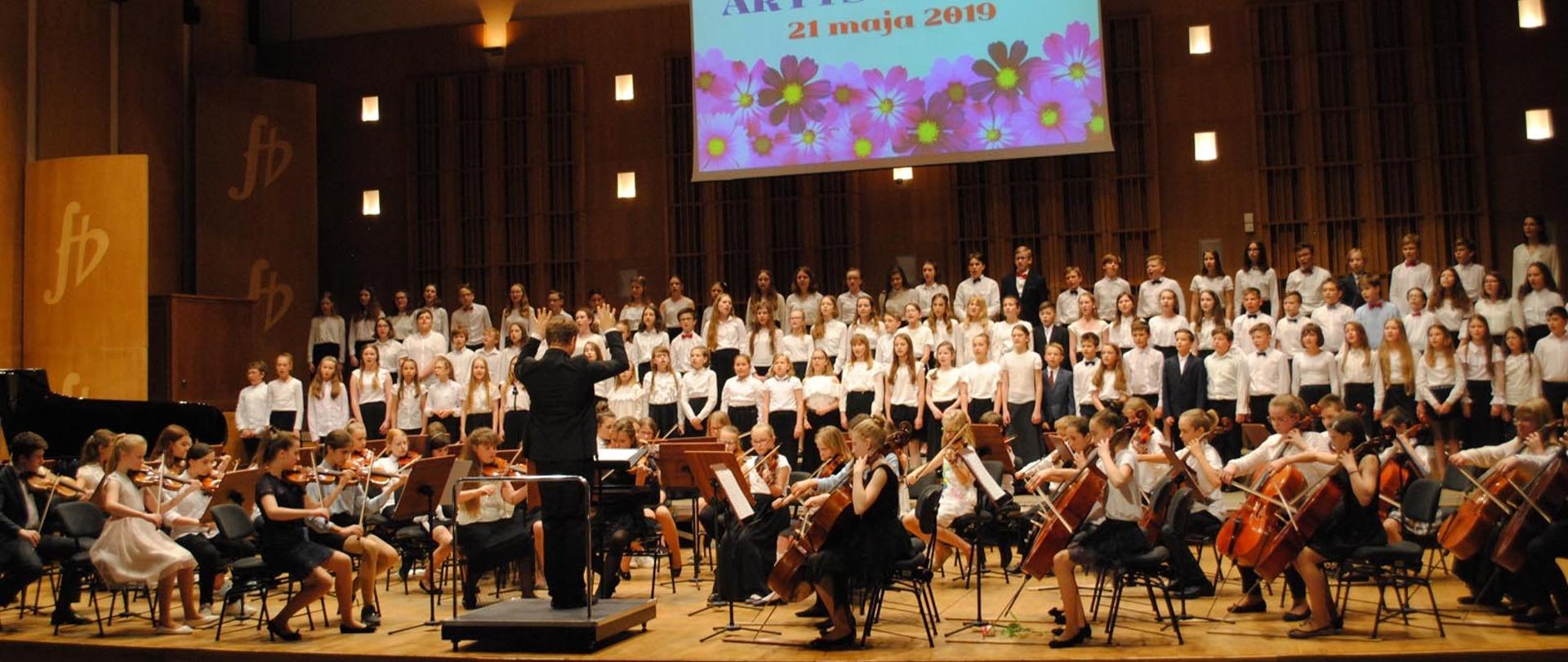 Kolorowe zdjęcie przedstawiające stojących w trzech rzędach uczniów Chóru PSM I stopnia oraz siedzących uczniów Orkiestry smyczkowej PSM I stopnia grających na skrzypcach i wiolonczelach. Na środku sceny znajduje się dyrygent całego zespołu. Wszyscy uczniowie ubrani są w białe bluzki oraz czarne spódnice lub spodnie. Ponadto na środku sceny znajduje się wiszący pod sufitem ekran na którym wyświetlona jest informacja: „Wiosna Młodzieży Artystycznej 21 maja 2019 r.”.