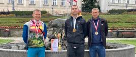Na zdjęciu widać trzech funkcjonariuszy z medalami oraz pucharem za zawody. Zdjęcie wykonane w dzień w parku przy fontannie. Panowie ubrani w ubrania cywilne. 