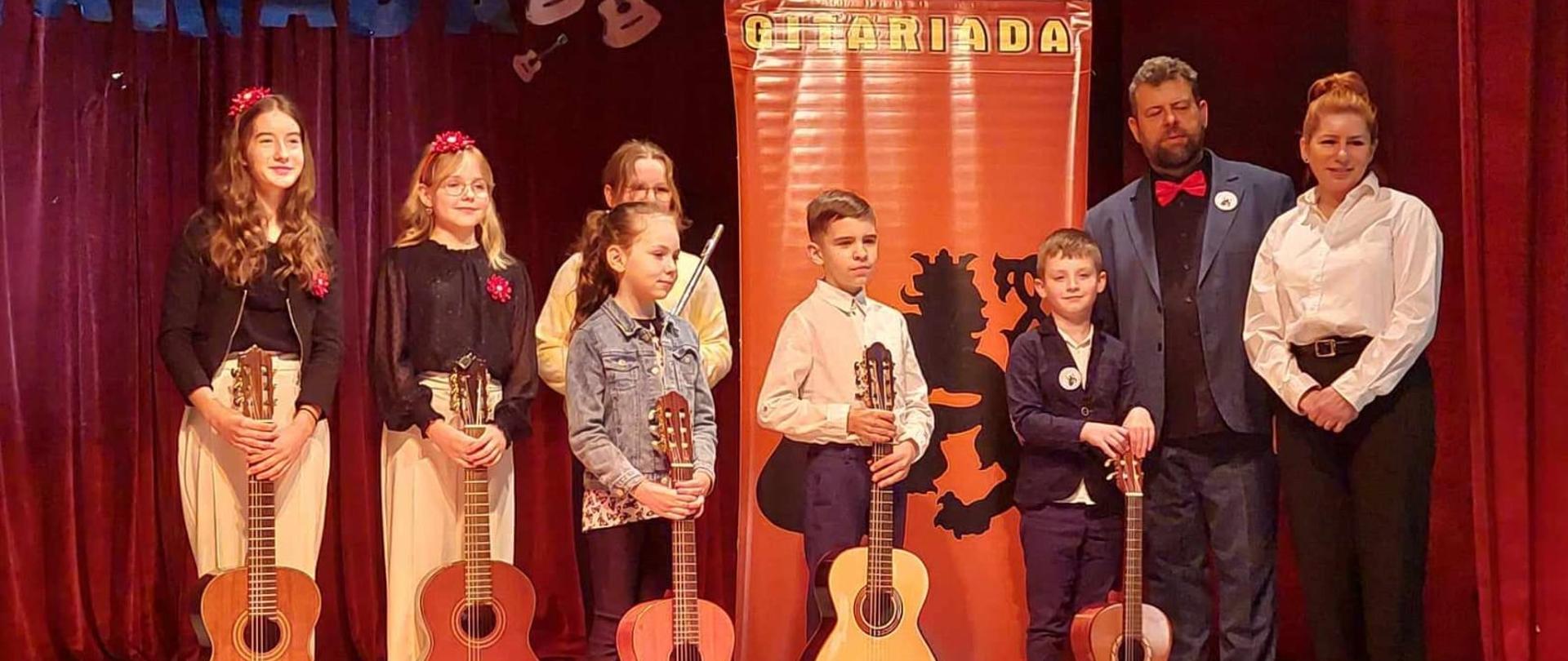 Grupa uczniów wraz z nauczycielem i kobietą stoją pozując do zdjęcia trzymając gitary na scenie