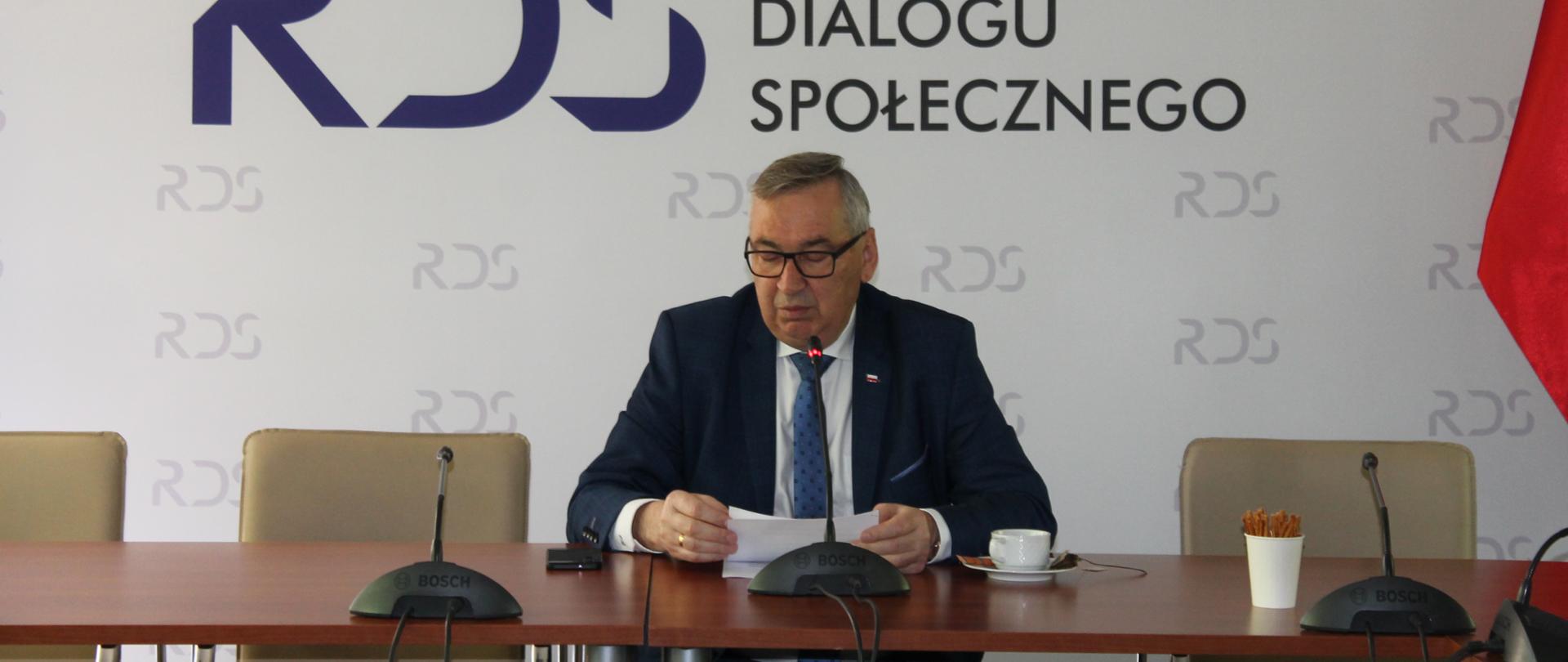 Na zdjęciu widnieje stół prezydialny za którym siedzi wiceminister Rodziny i polityki społecznej – Stanisław Szwed, który prowadzi spotkanie. W tle znajduje się baner z napisem Rada Dialogu Społecznego.