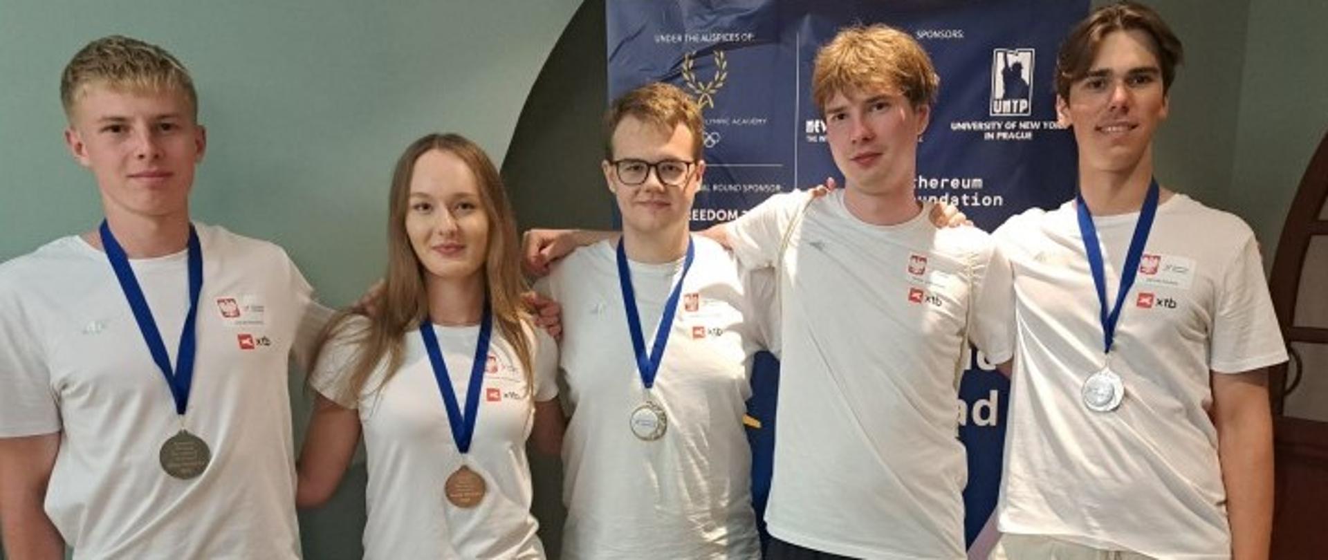 Pięcioro młodych ludzi stoi w rzędzie, uśmiechając się do kamery. Wszyscy są ubrani w białe koszulki z małym, czerwonym logo na piersi i mają na szyi medale na niebieskich wstążkach. Pośrodku stoi kobieta, a wokół niej są czterej mężczyźni. Dwoje młodych ludzi z prawej strony opiera ręce na ramionach pozostałych. W tle widoczny jest baner z logami sponsorów i napisami w języku angielskim.