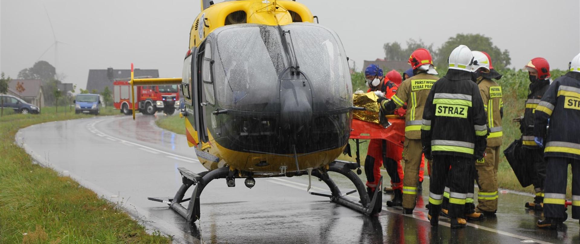 Stojący na drodze śmigłowiec Lotniczego Pogotowia Ratunkowego oraz ratownicy medyczni i strażacy transportujący poszkodowanego. W tle samochód strażacki oraz zabudowania.