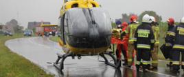 Stojący na drodze śmigłowiec Lotniczego Pogotowia Ratunkowego oraz ratownicy medyczni i strażacy transportujący poszkodowanego. W tle samochód strażacki oraz zabudowania.