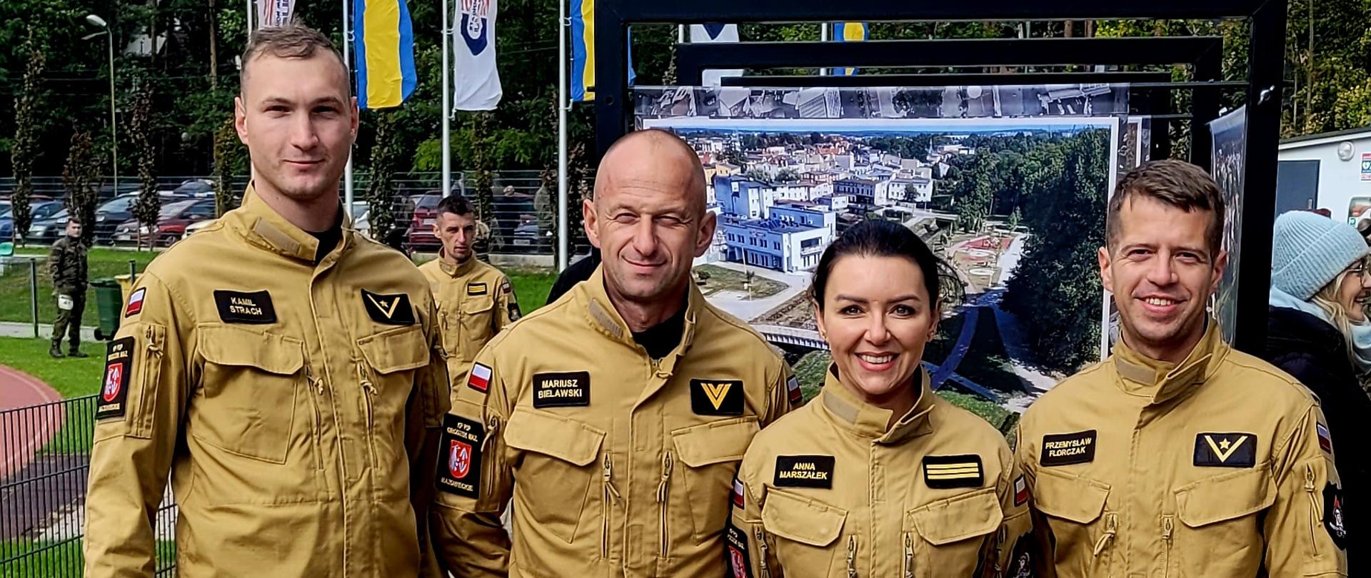 Grupowe zdjęcie czterech osób – trzech mężczyzn i jednej kobiety – stojących na wybrukowanym placu. Wszyscy są ubrani w beżowe kombinezony z ciemnymi pagonami i godłami na ramionach. Noszą plakietki startowe z numerami. Stoją przed dużym panelem wystawowym z fotografią przedstawiającą widok fabryki lub dużego obiektu przemysłowego. Za nimi widoczne są maszty z flagami.