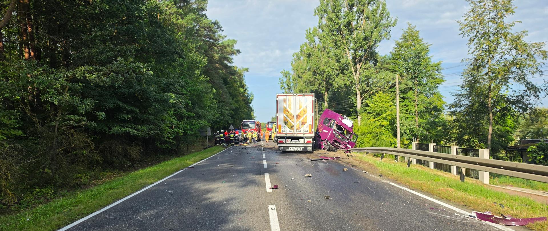 Droga asfaltowa, odcinek leśny. Po prawej stronie widoczny TIR z rozbitą fioletową kabiną (ciągnikiem) częściowo znajduje się w rowie, naczepa częściowo na poboczu. Z lewej strony widoczni funkcjonariusze udzielający pomocy osobie znajdującej się w rozbitym samochodzie osobowym. W tle czerwone samochody bojowe PSP i OSP. 