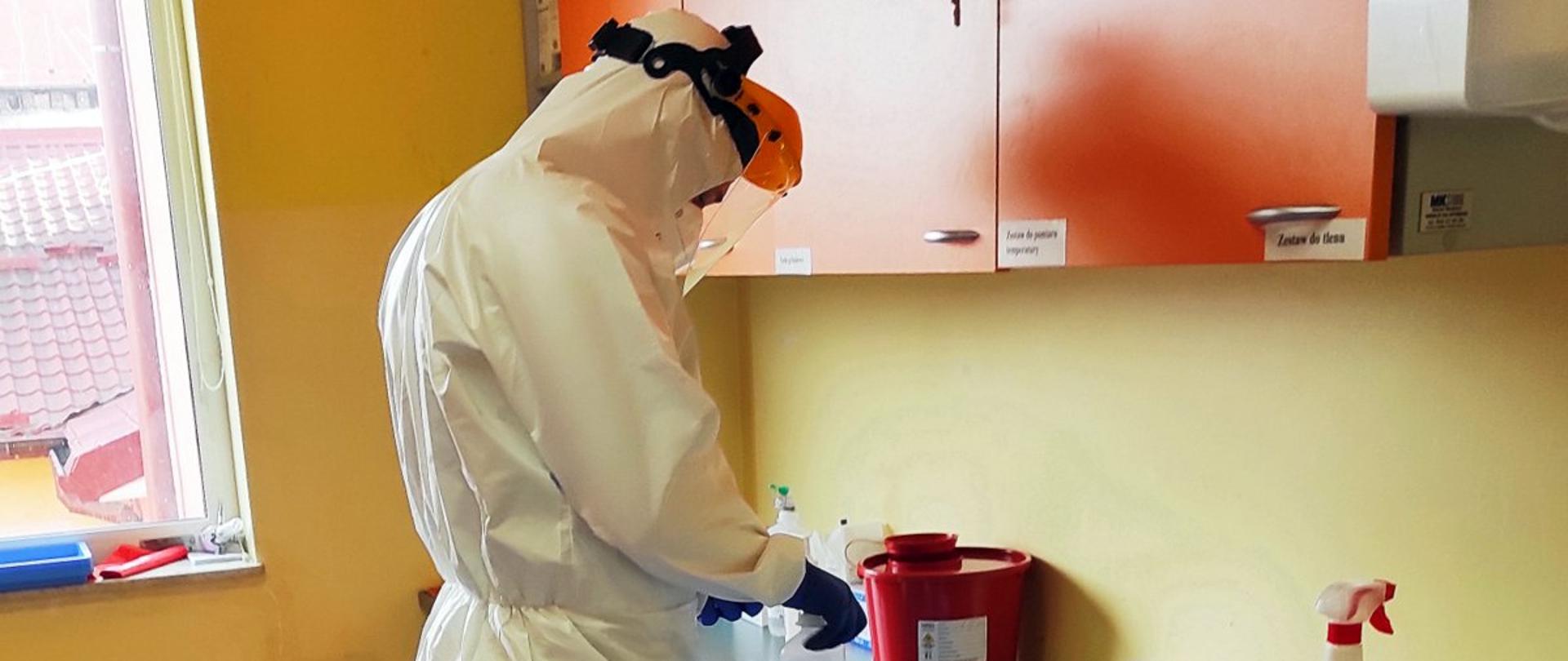 Na zdjęciu osoba ubrana w biały strój ochronny, przyłbicę i gumowe rękawiczki pracuje przy stole w gabinecie zabiegowym.