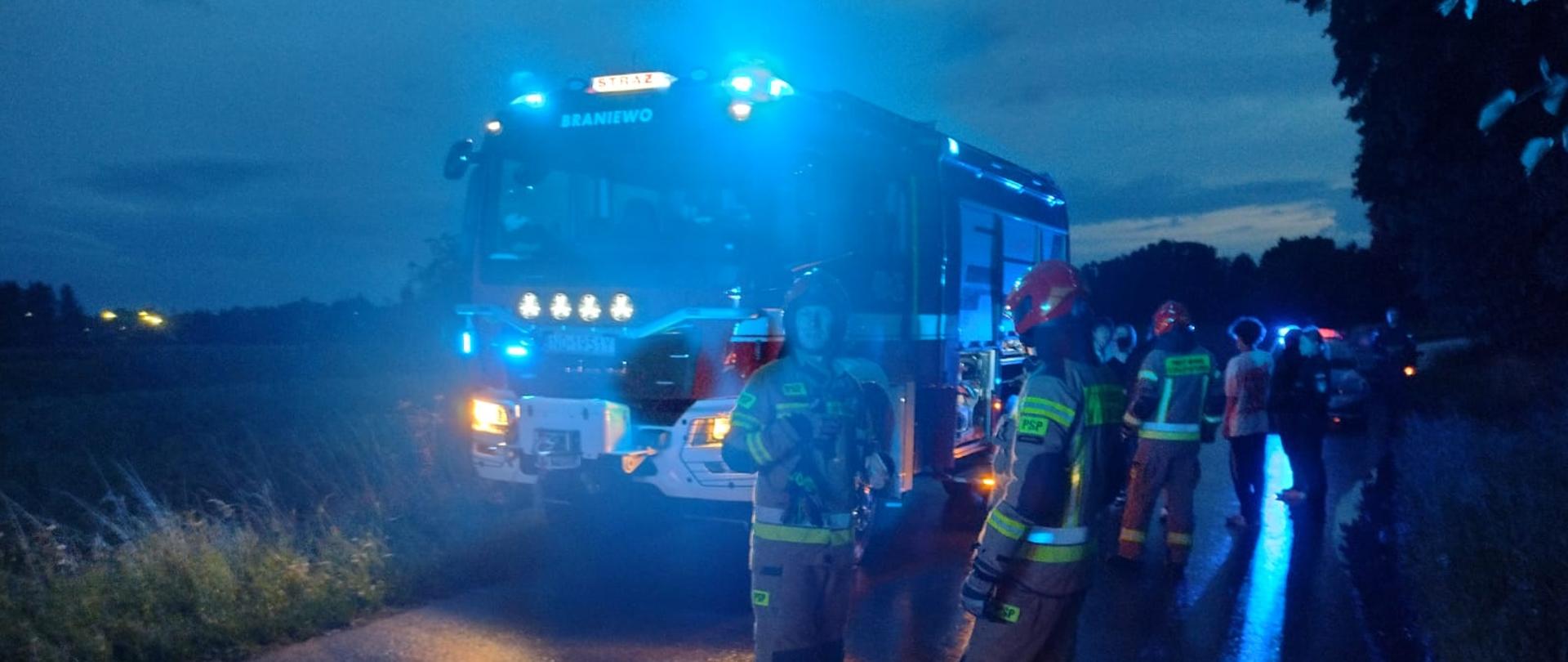 Zdjęcie zrobione nocą. na drodze ustawiony samochód strażacki na niebieskich światłach ostrzegawczych i strażacy w mundurach i czerwonych hełmach.