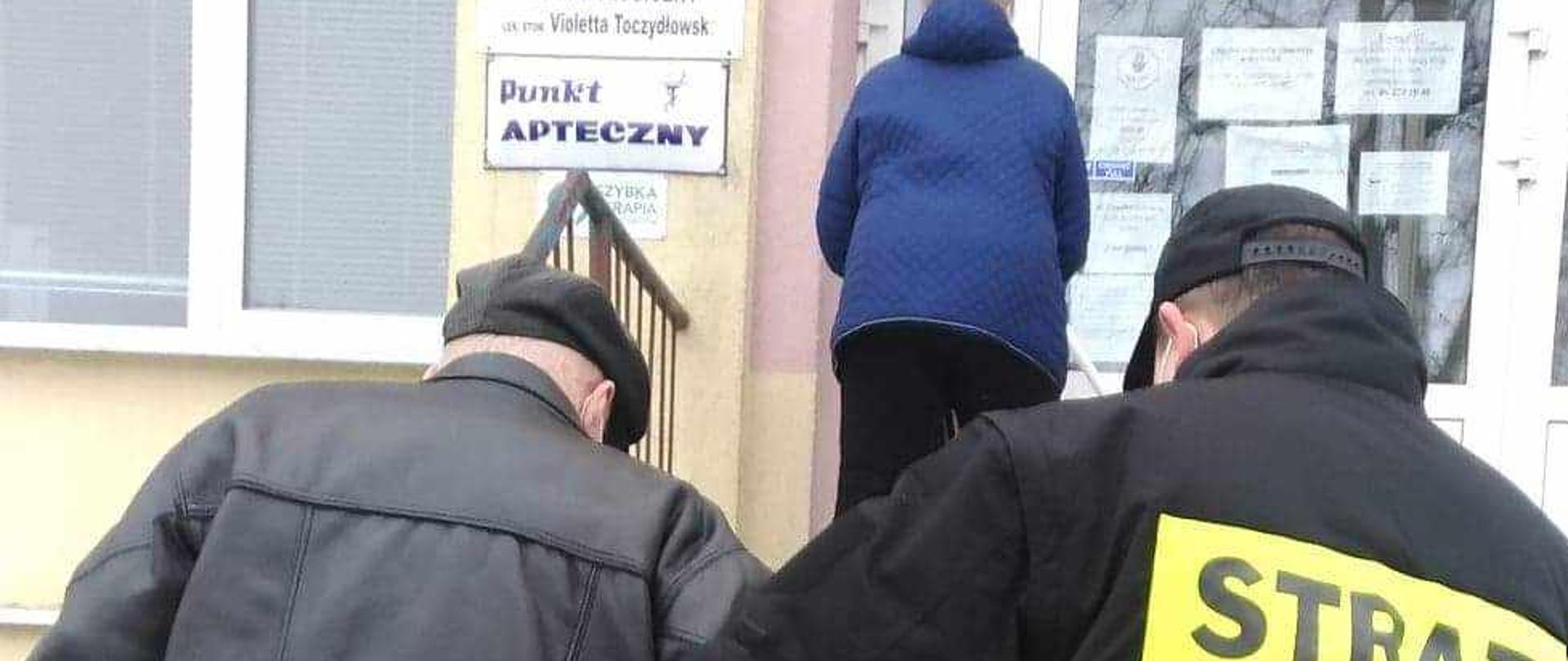 Na zdjęciu strażak OSP w ubraniu koszarowym pomaga starszej osobie wejść po schodach do przychodni NFZ.