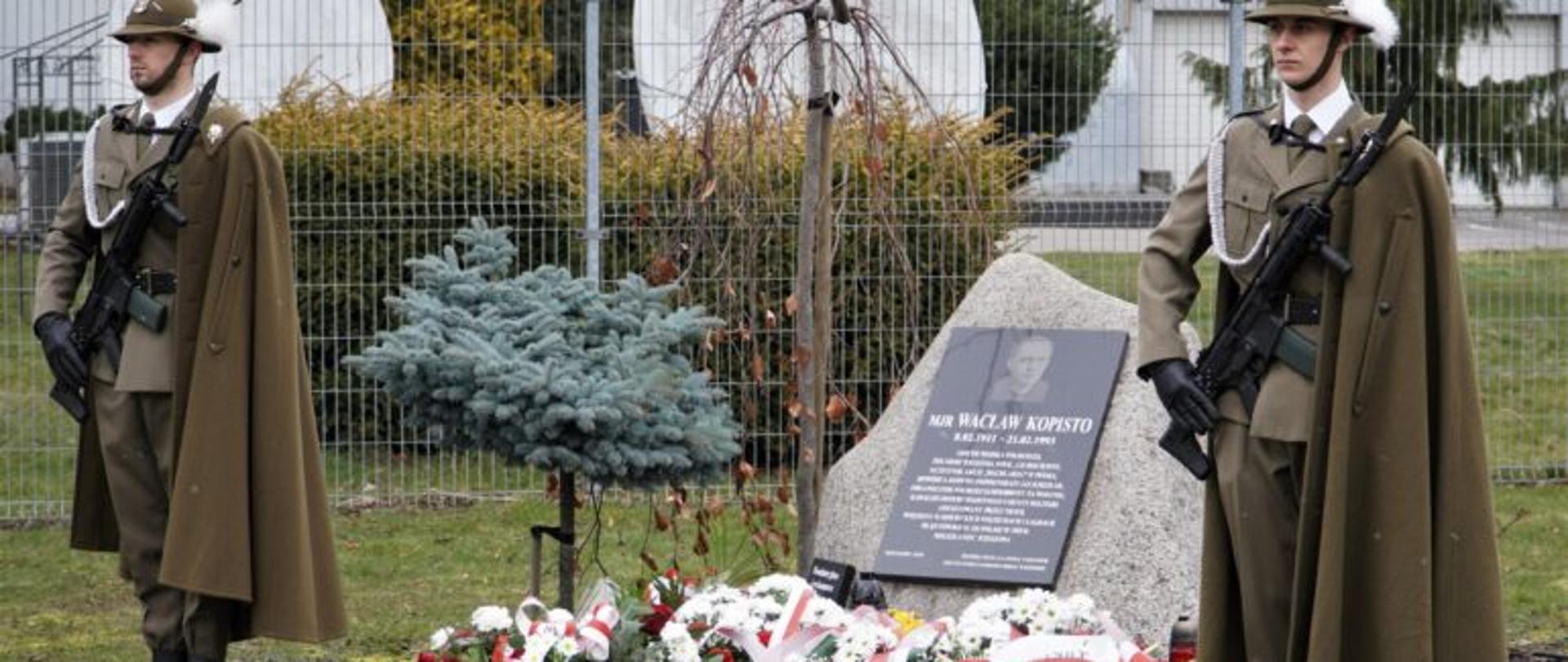 Żołnierze stojący obok tablicy upamiętniającej śp. mjr. Wacława Kopisto. Przed tablicą znajdują się wiązanki kwiatów wieńce.