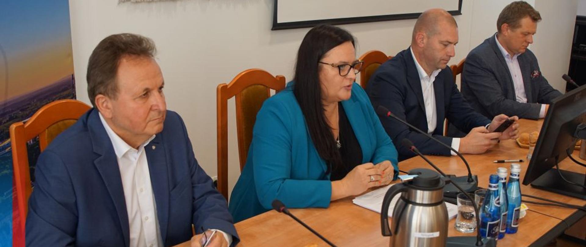 Trzech mężczyzn i wiceminister Małgorzata Jarosińska-Jedynak pomiędzy nimi siedzą przy stole. Na nim monitor, mikrofony i butelki.