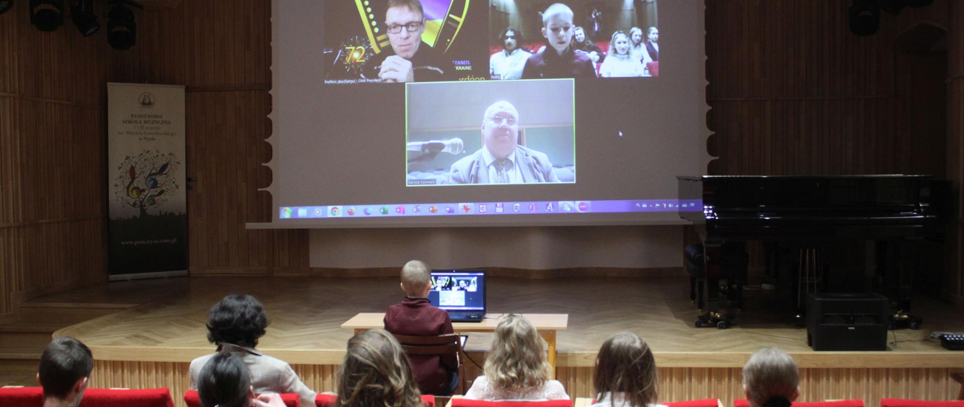 Zdjęcie. Spotkanie online uczniów z nauczycielami. Na pierwszym planie widać od tyłu słuchaczy biorących udział w spotkaniu. Dalej, przed estrada stoi ławka w laptopem, przy którym siedzi osoba zadająca pytanie. W tle na wiszącym nad estradą dużym ekranie widać trzy okienka, w których pojawiają się osoby biorące udział w dyskusji.