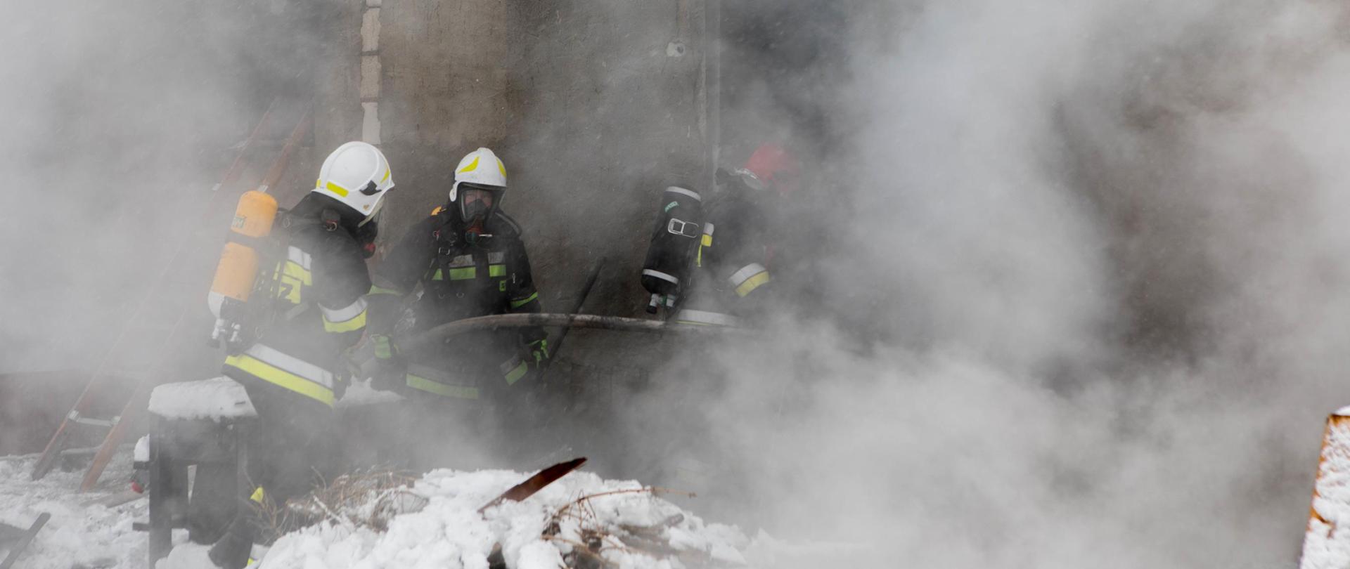 Na zdjęciu widnieją strażacy w trakcie prowadzenia działań gaśniczych przy pożarze warsztatu samochodowego. Jest dzień, słonecznie, są opady śniegu. 