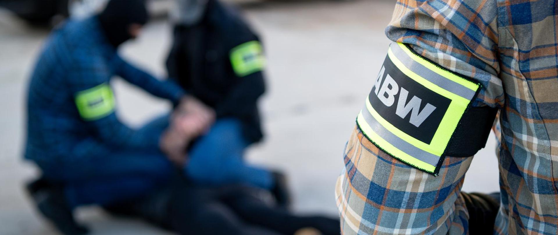Po prawej stronie zdjęcia widać ramię funkcjonariusza ABW. Funkcjonariusz jest ubrany w koszulę w kratkę (kolory: szary, niebieski, biały, brązowy i pomarańczowy). Na przedramieniu funkcjonariusza widać odblaskowe oznaczenie z napisem ABW. Po lewej stronie zdjęcia widać dwóch funkcjonariuszy ABW w kominiarkach i z odblaskowymi oznaczeniami, zatrzymujących osobę. Osoba zatrzymana leży na ziemi i są jej zakładane kajdanki. U góry zdjęcia widać również fragment ciemnego samochodu.