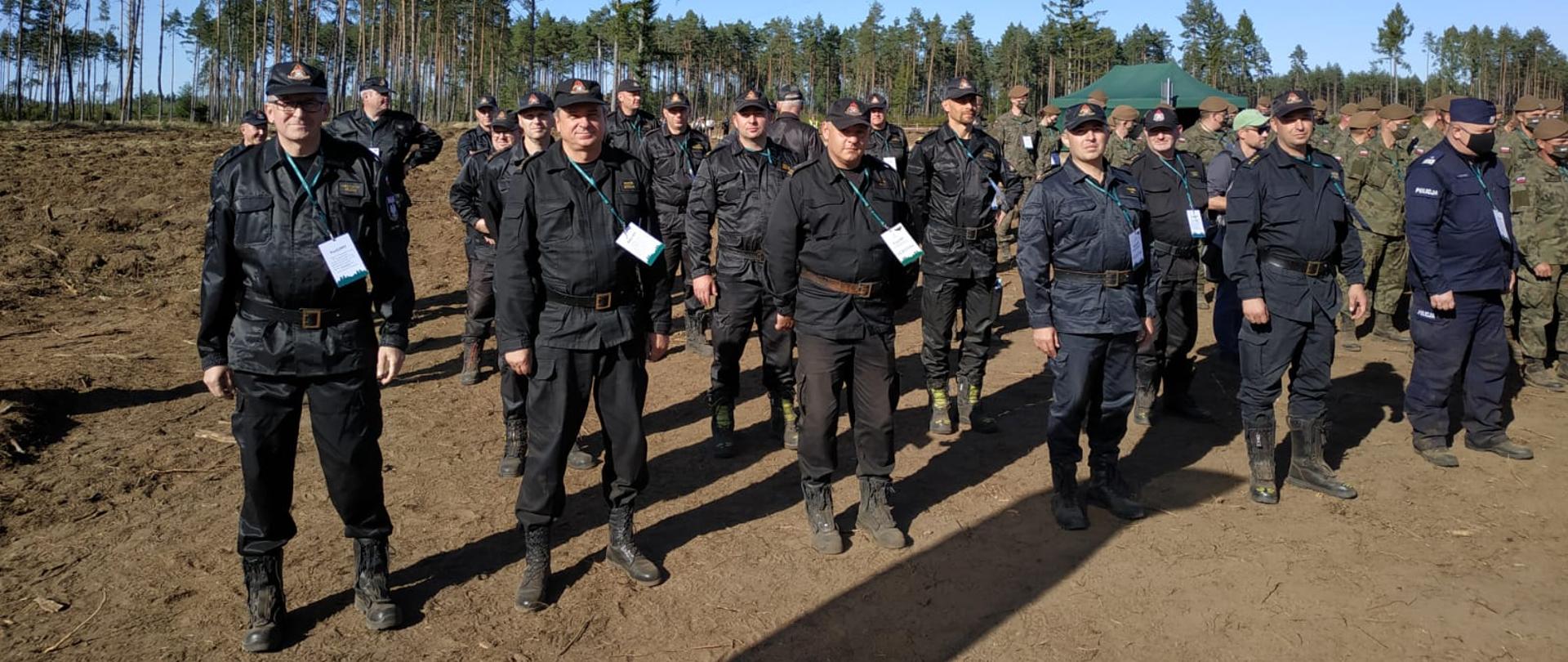 Zdjęcie przedstawia grupę mężczyzn stojących w czterech rzędach na tle lasu.