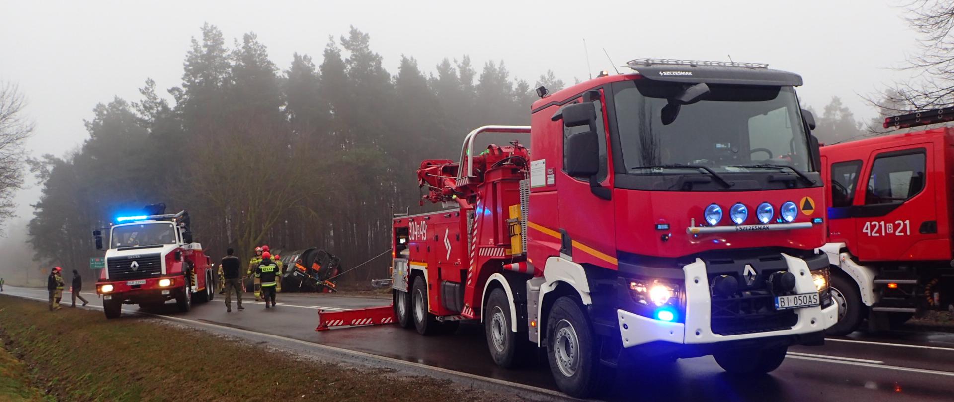 2 ciężkie samochody ratownictwa technicznego typu Mega City stoją na drodze. W tle przewrócona cysterna. Dookoła pracują strażacy i żołnierze.