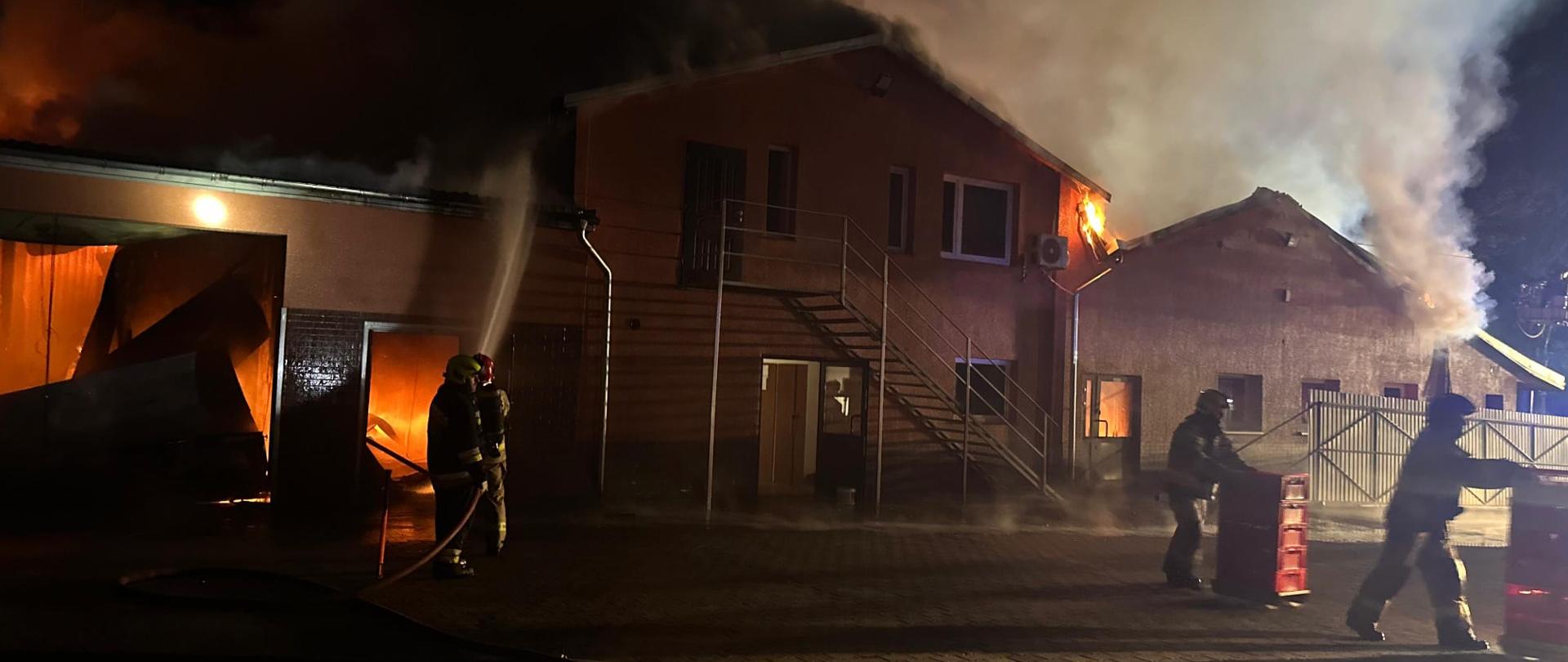 Strażacy prowadzą działania gaśnicze przy budynku objętym pożarem – jeden z nich kieruje strumień wody do wnętrza, podczas gdy z dachu i okien wydobywa się dym i ogień