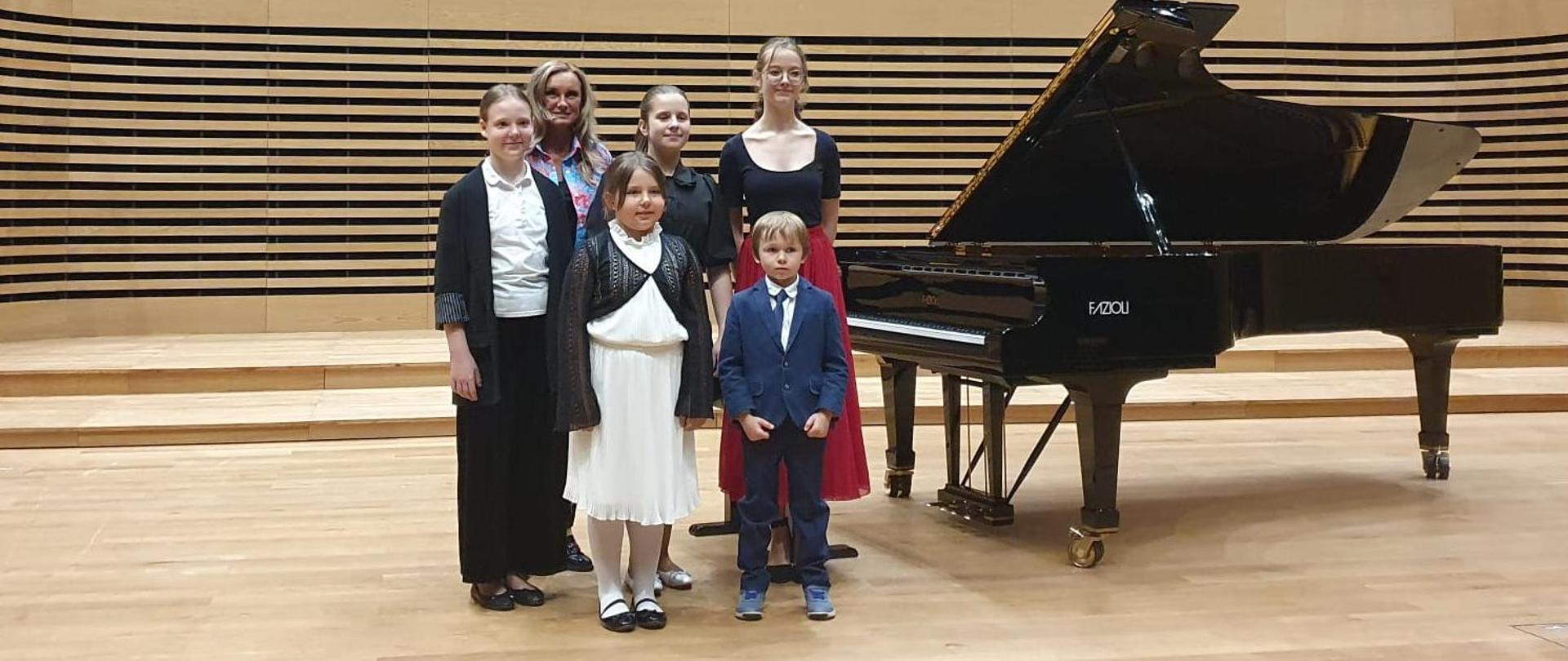 Kobieta, mały chłopczyk i dziewczynka i trzy starsze dziewczyny stoją przy fortepianie Fazioli na estradzie sali koncertowej.