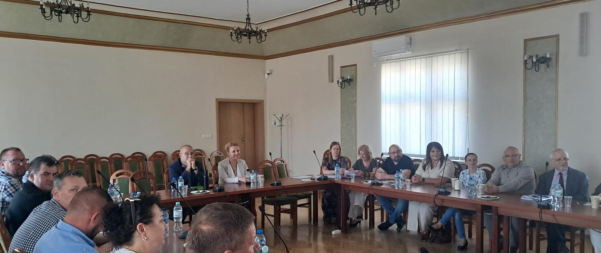 Zdjęcie przedstawia spotkanie kilkunastu osób w sali konferencyjnej. Uczestnicy siedzą przy drewnianych stołach ustawionych w kształt podkowy. Na stołach znajdują się mikrofony, butelki z wodą oraz dokumenty. W tle widoczne są krzesła, duże okno z pionowymi żaluzjami oraz dekoracyjne żyrandole. Część uczestników patrzy w kierunku osoby wykonującej zdjęcie.