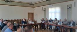 Zdjęcie przedstawia spotkanie kilkunastu osób w sali konferencyjnej. Uczestnicy siedzą przy drewnianych stołach ustawionych w kształt podkowy. Na stołach znajdują się mikrofony, butelki z wodą oraz dokumenty. W tle widoczne są krzesła, duże okno z pionowymi żaluzjami oraz dekoracyjne żyrandole. Część uczestników patrzy w kierunku osoby wykonującej zdjęcie.