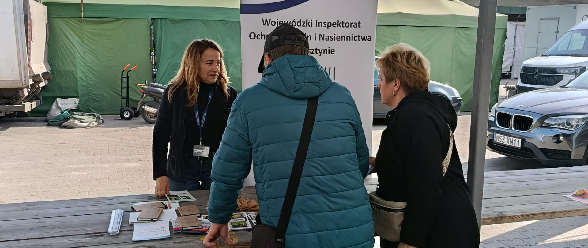 Kobieta stoi przy rollupie WIORiN naprzeciwko pochylonych do niej osób, pomiędzy nimi stół na którym leżą ulotki