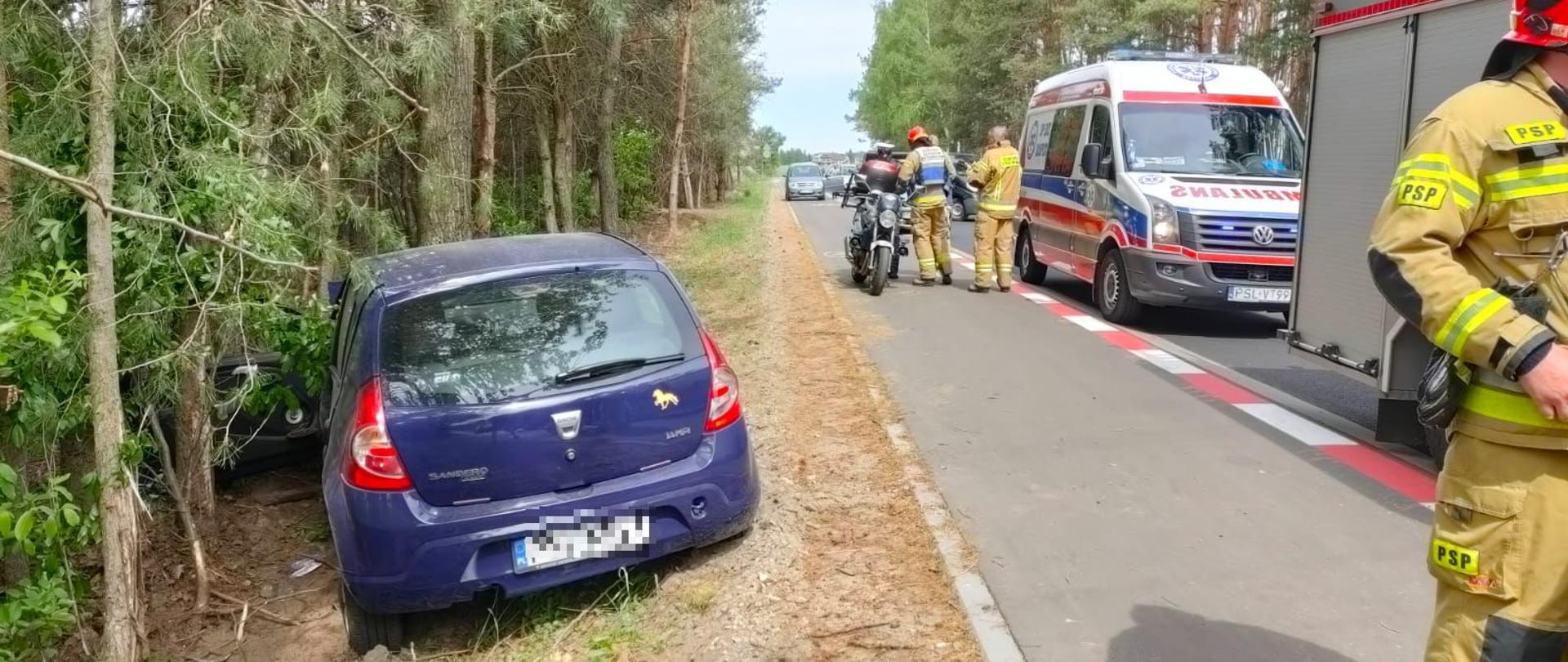 niebieski samochód osobowy na poboczu drogi, który uderzył w drzewo na drodze widać karetkę pogotowia, samochód strażacki oraz motor policji w oddali na drodze inne samochody wokół drzewa