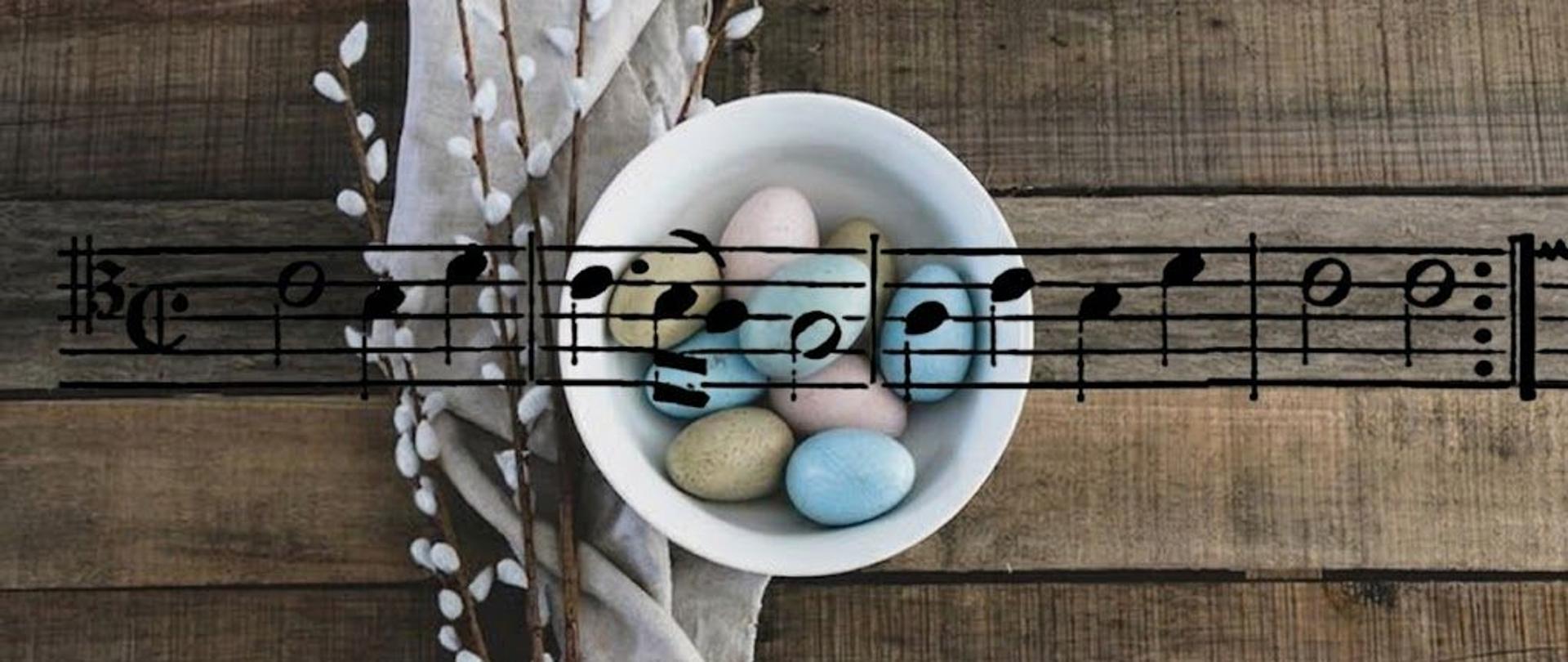 Grafika kolorowa. Tło obrazu stanowią ciemnobrązowe, poziome deski o wyraźnej strukturze drewna. Po lewej stronie widoczny jest pionowy fragment jasnej, lnianej tkaniny, obok której leżą gałązki bazi. W centrum znajduje się biała, okrągła miseczka wypełniona kilkoma pisankami w pastelowych kolorach (błękitnym, beżowym i różowym). Przez środek całego obrazu przebiega czarny, lekko półprzezroczysty fragment zapisu nutowego w kluczu altowym, który nakłada się bezpośrednio na miseczkę z jajkami i drewniane tło.