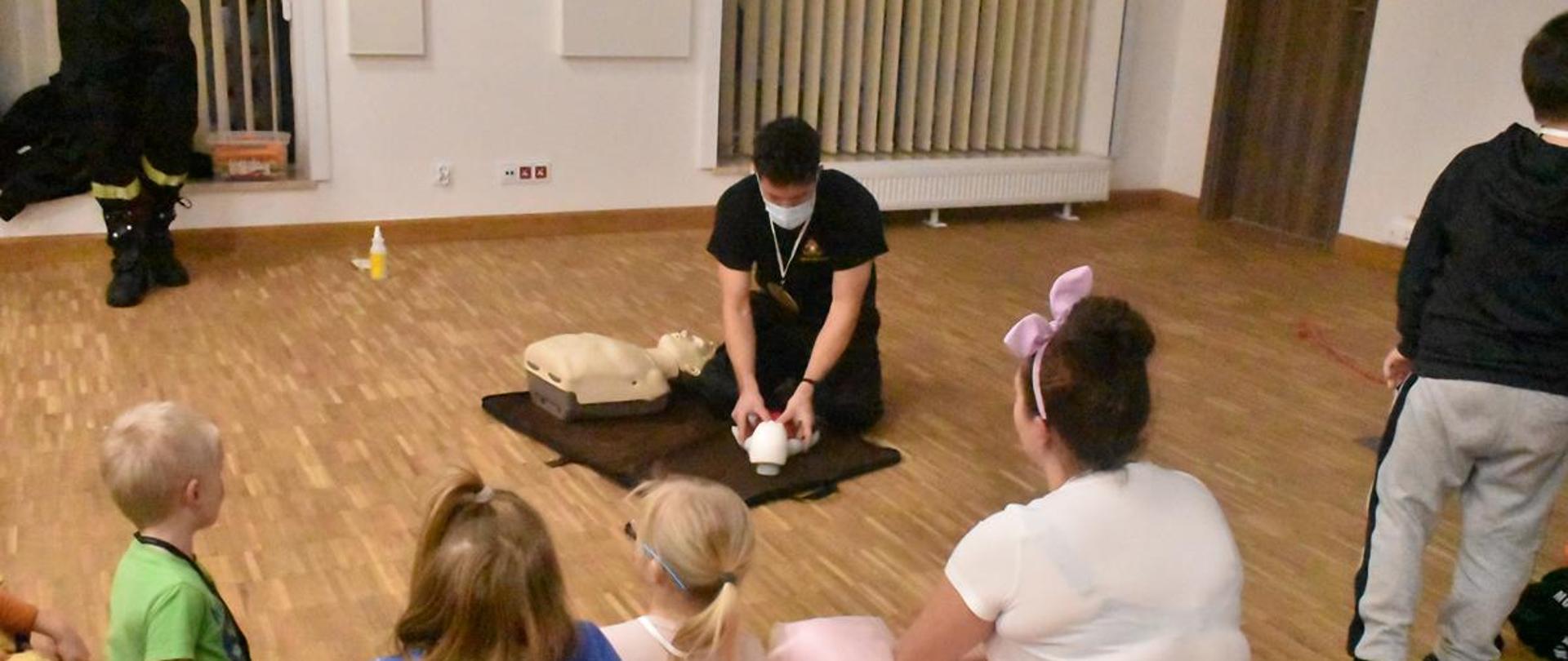 Strażak podczas pokazu udzielania pierwszej pomocy. Strażak demonstruje wykonywanie resuscytacji krążeniowo-oddechowej na manekinie niemowlaka. Przed nim siedzą dzieci.