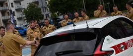 Grupa strażaków w umundurowaniu służbowym stoi na zewnątrz wokół białego samochodu elektrycznego Nissan Leaf oznakowanego logo Państwowej Straży Pożarnej. Prowadzący omawia uczestnikom zasady postępowania podczas działań ratowniczych przy pojeździe elektrycznym.