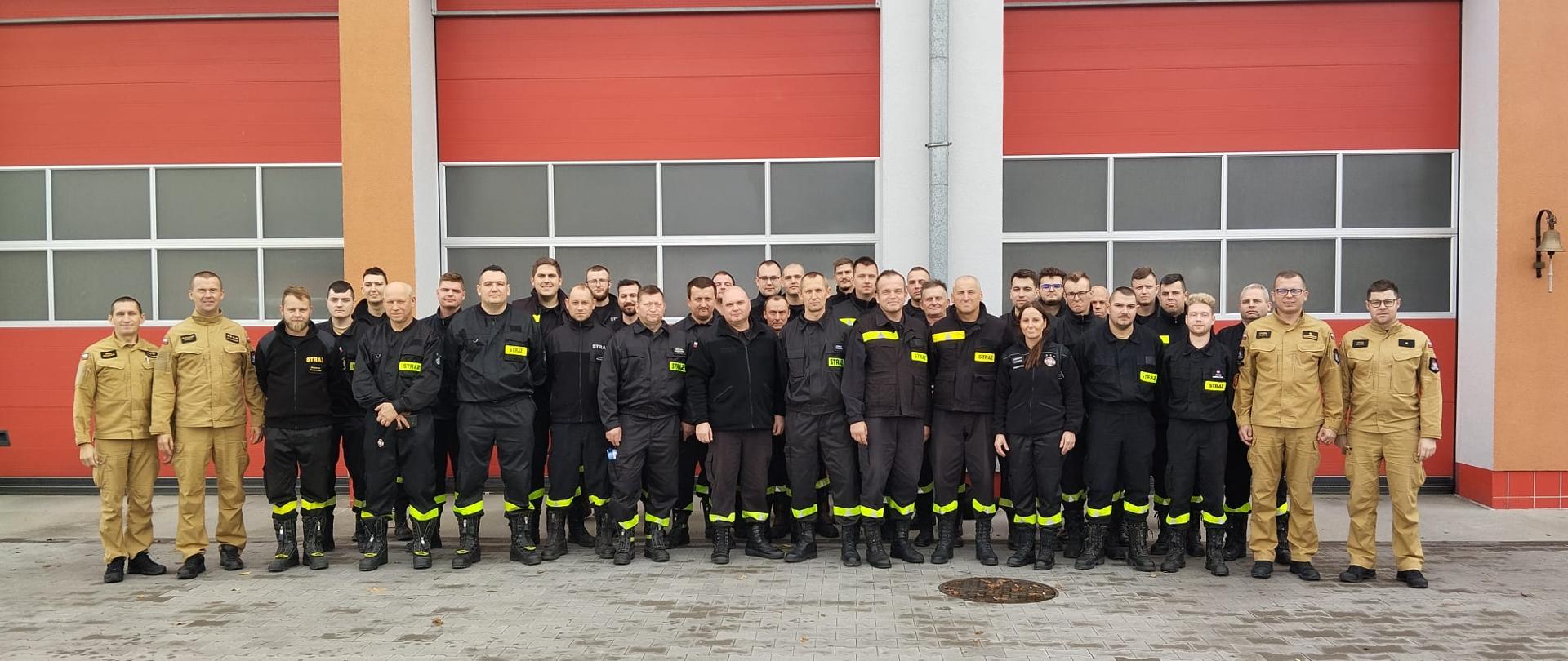 Strażący w mundurach stoją w dwuszeregu na tle bram garażowych.