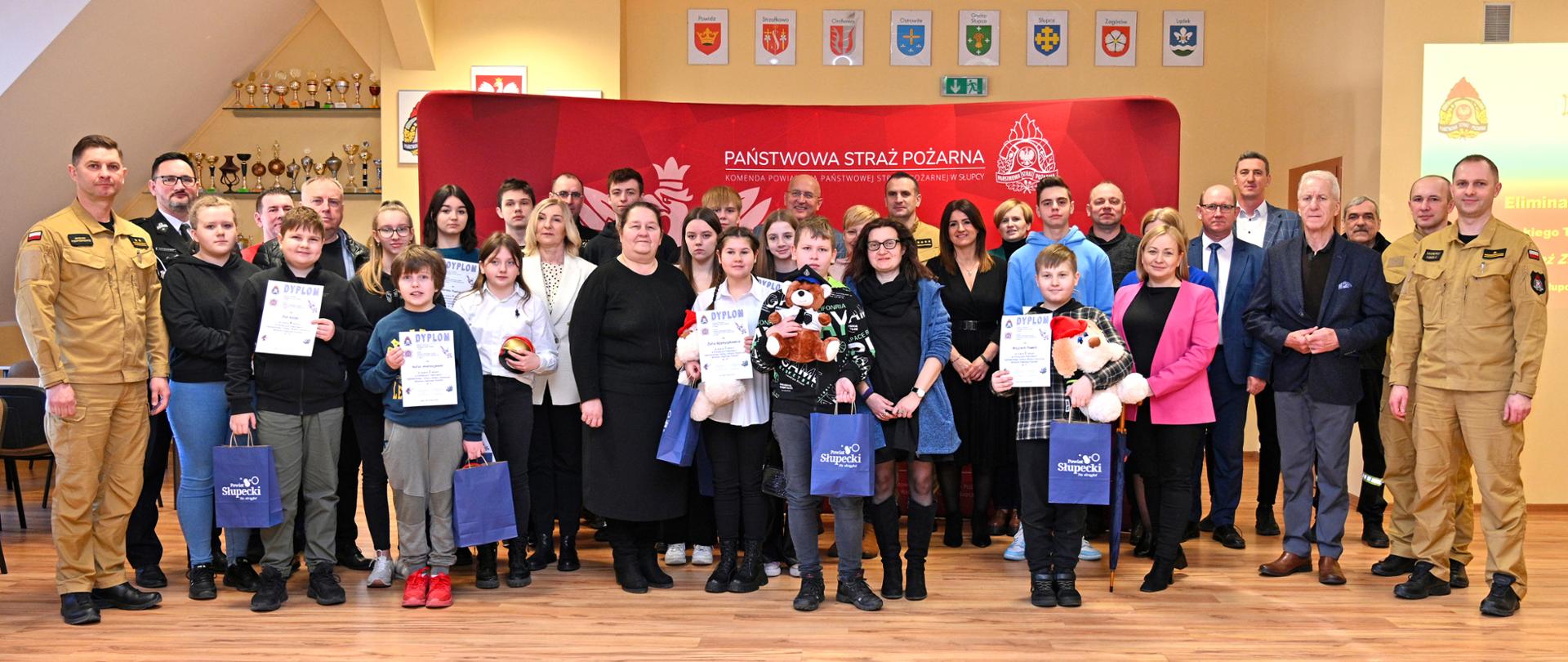 Laureaci, opiekunowie turnieju oraz przedstawiciele straży pożarnej na tle ścianki z koloru czerwonego w obecności strażaków oraz inne osoby