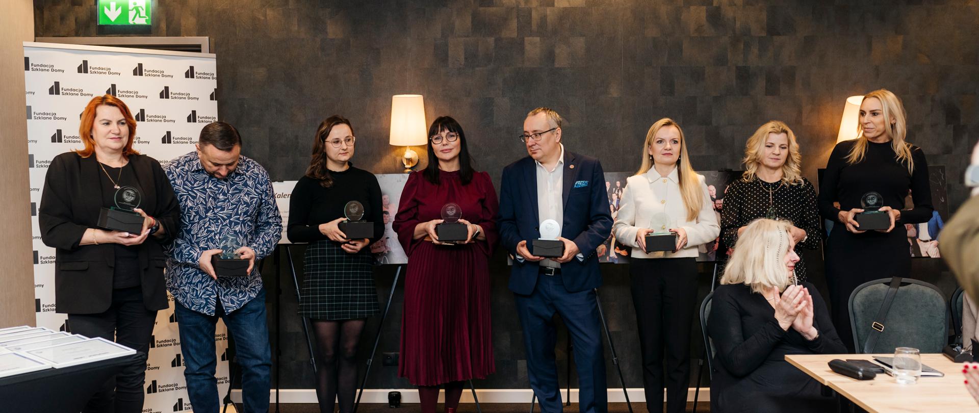 Grupa osób, kobiety i mężczyźni pozują do zdjęcia. W dłoniach trzymają statuetkę. Pozostałe osoby widoczne na zdjęciu klaszczą w dłonie. 