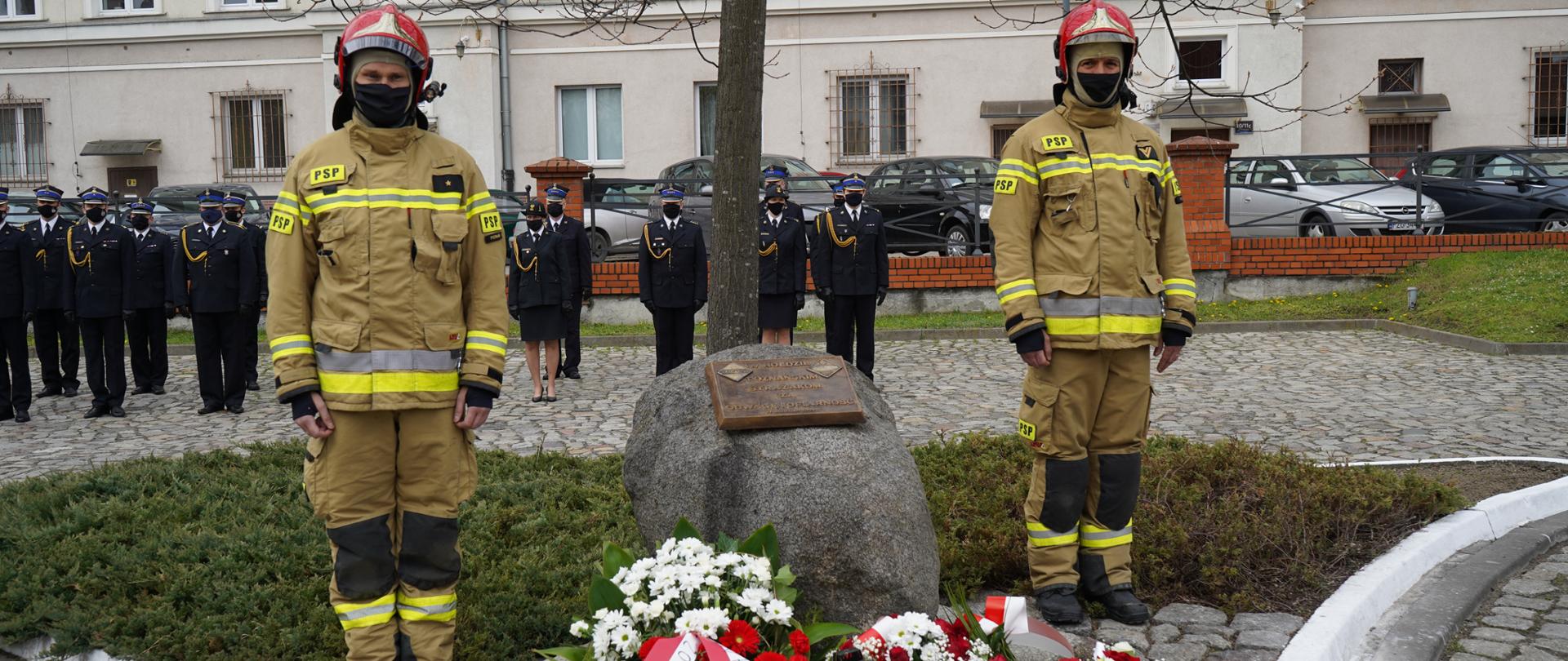 Przed kamieniem - pomnikiem stoi dwóch strażaków w ubraniach specjalnych, przed kamieniem leżą wiązanki z kwiatami oraz znicz. W tle kadra keirownicza strażaków
