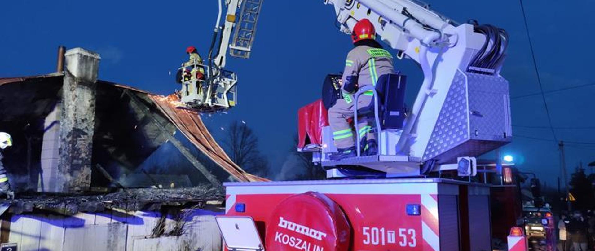 Strażacy pracują w koszu drabiny mechanicznej, usuwają arkusze blachy z spalonej konstrukcji dachu.
