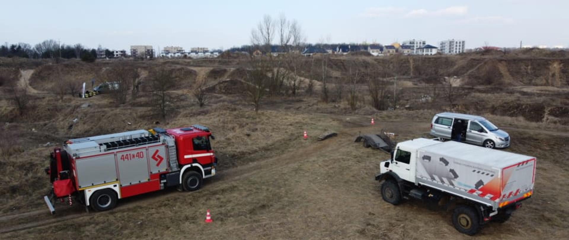 Widok z góry na pagórkowaty, piaszczysty, pokryty suchą trawą teren, na którym stoją pojazdy. Po lewej czerwono - srebrny, ciężarowy samochód strażacki. Po prawej biały terenowy samochód ciężarowy i srebrny dostawczy. W tle drzewa i krzewy, a na dalszym planie budynki osiedla mieszkaniowego.