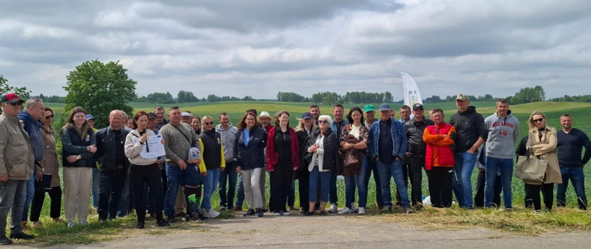 duża grupa osób pozuje do zdjęcia, za nimi pola uprawne, przed nimi droga asfaltowa