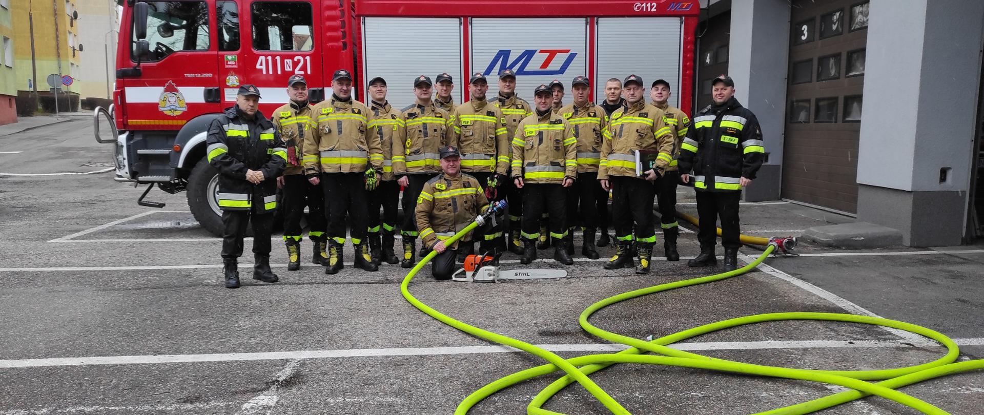 Na tle dużego czerwonego samochodu strażackiego. Duża grupa strażaków w mundurach pozuje do zdjęcia. Rozwinięte zielone węże strażackie.