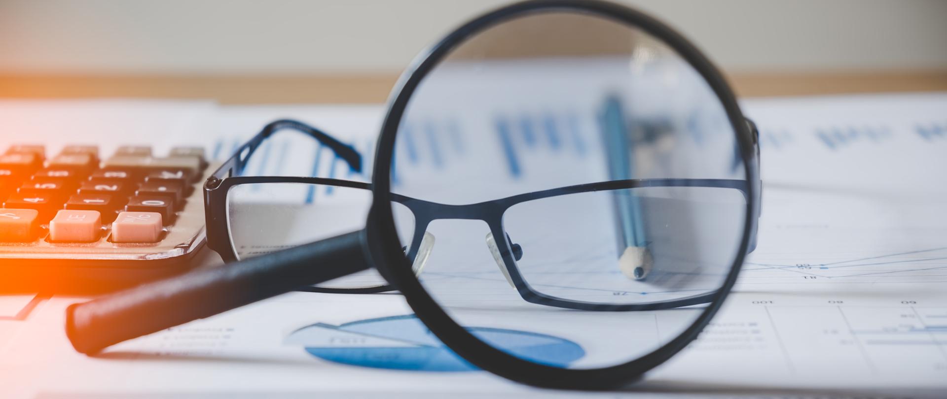 Wykresy i tabele pod szkłem powiększającym, obok ołówek i kalkulator