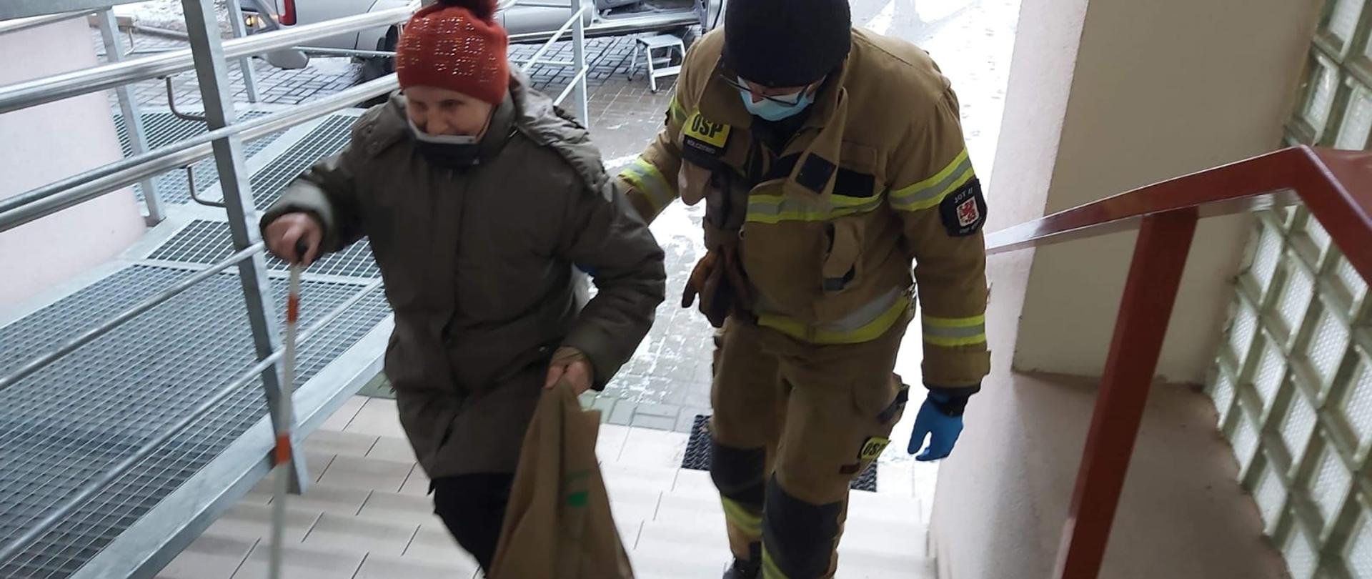 Zdjęcie przedstawia strażaka który pomaga wejść starszej osobie do punktu szczepień.