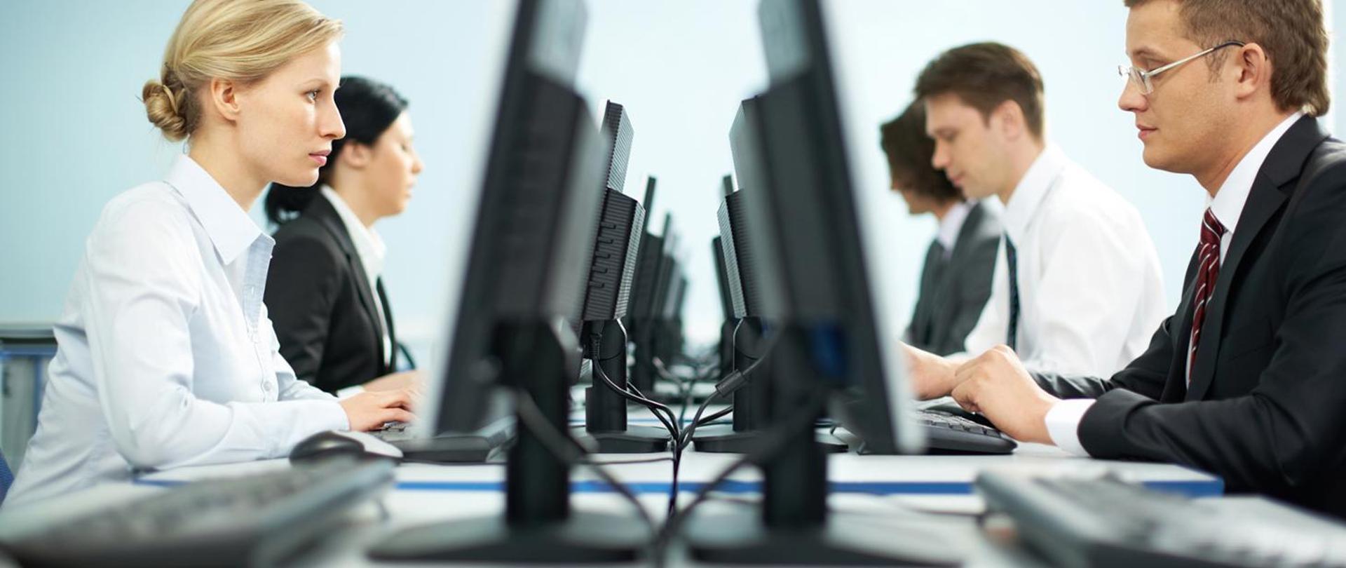 na zdjęciu stół z monitorami komputerów skierowanymi w przeciwne strony przed którymi siedzą pracownicy