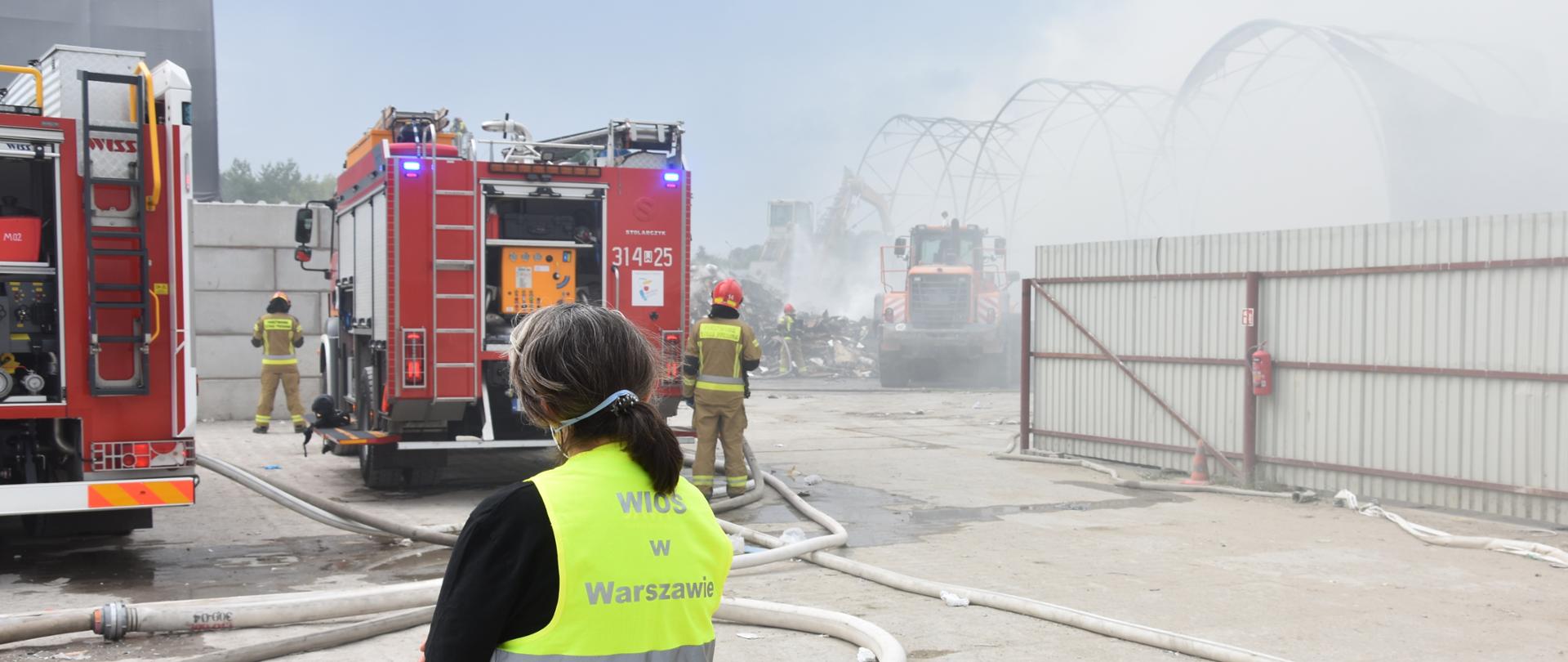 Inspektor Wojewódzkiego Inspektoratu Ochrony Środowiska w Warszawie prowadzi działania na miejscu pożaru. W tle po lewej stronie widać dwa wozy bojowe Państwowej Straży Pożarnej. Po prawej stronie, za stalowym, szarym płotem o wysokości 2 metrów widać ciągnik oraz stalowe konstrukcje spowite szarym dymem.