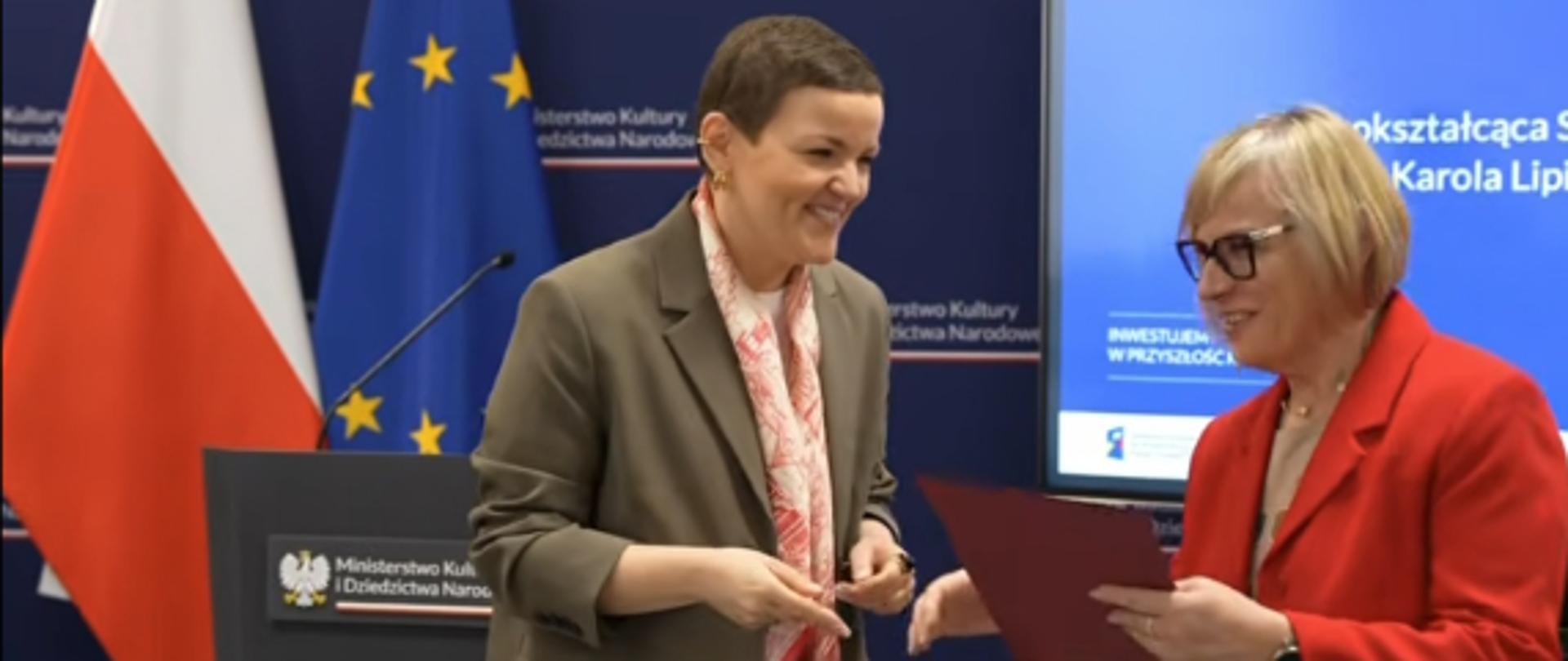 Zdjęcie przedstawia sylwetki dwóch osób: Panią Minister Kultury i Dziedzictwa Narodowego Martę Cienkowską wręczającą dyplom z gratulacjami Pani Dyrektor Ogólnokształcącej Szkoły Muzycznej I i II stopnia im. Karola Lipińskiego w Lublinie Beacie Syczuk. W tle widoczna jest Flaga Narodowa Polski, Flaga Unii Europejskiej, logo Ministerstwa Kultury i Dziedzictwa Narodowego oraz część ekranu telewizyjnego wyświetlającego napis Ogólnokształcąca Szkoła Muzyczna I i II stopnia im. Karola Lipińskiego w Lublinie.