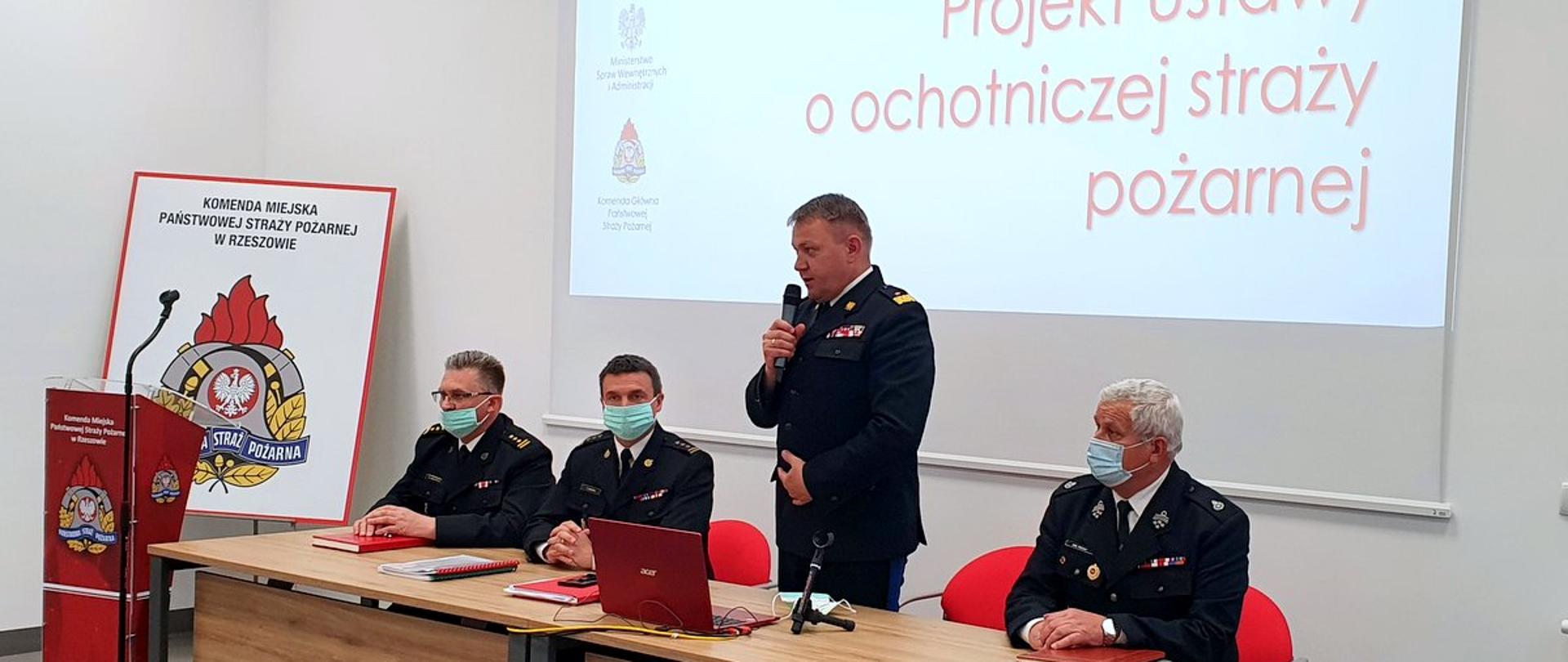 Zdjęcie przedstawia uczestników spotkania konsultacyjnego w sprawie projektu Ustawy o ochotniczej straży pożarnej na świetlicy Jednostki Ratowniczo-Gaśniczej Nr 3 w Rzeszowie. Centralne miejsca zajmuje podkarpacki komendant wojewódzki, komendant miejski PSP w Rzeszowie wraz ze swoim zastępcą oraz prezes zarządu powiatowego Związku Ochotniczych Straży Pożarnych Rzeczypospolitej Polskiej. Podkarpacki komendant wojewódzki przemawia do mikrofonu w pozycji stojącej. 