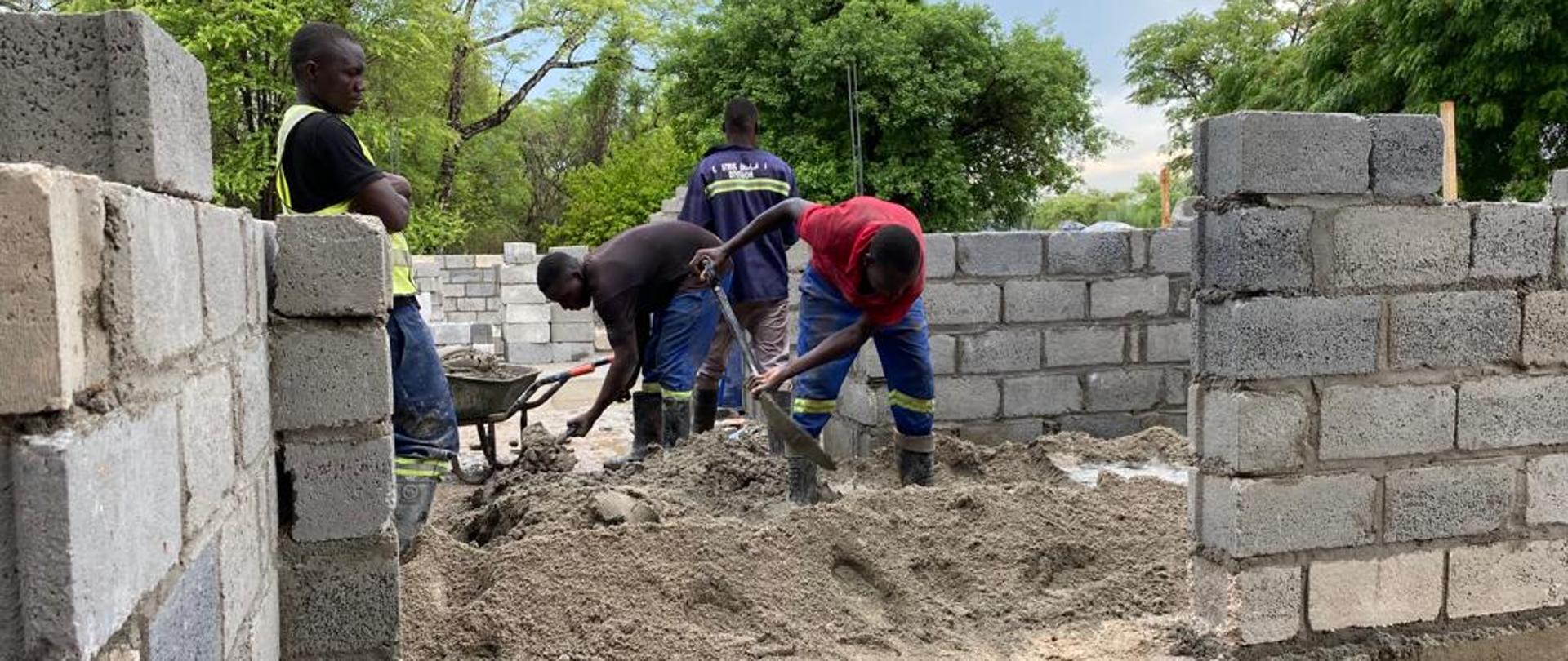 Grupa robotników pracuje na terenie budowy, przerzucając materiał budowlany przy użyciu łopat