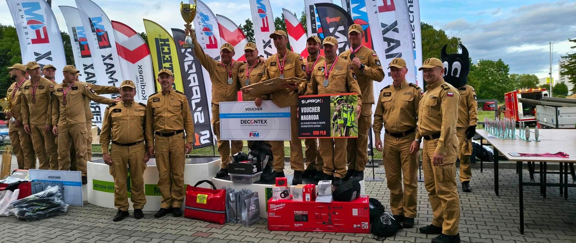 Grupa strażaków w beżowych mundurach stoi na podium podczas dekoracji po zawodach. Trzymają puchary, medale i nagrody rzeczowe, w tym voucher o wartości 500 zł. Za nimi stoją pionowe flagi sponsorów. Zdjęcie dokumentuje sukces reprezentacji województwa opolskiego — zdobycie tytułu wicemistrzów Polski w ratownictwie technicznym.