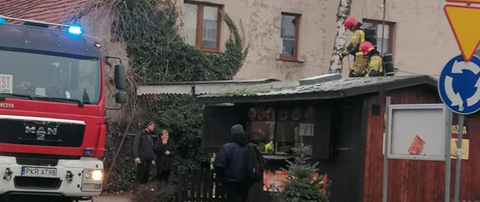 Zdjęcie przedstawia drewniany kiosk gastronomiczny, na dachu strażacy dokonują oceny stanu technicznego dachu, na drodze stoi pojazd straży pożarnej, a przed budynkiem stoją użytkownicy obiektu. 